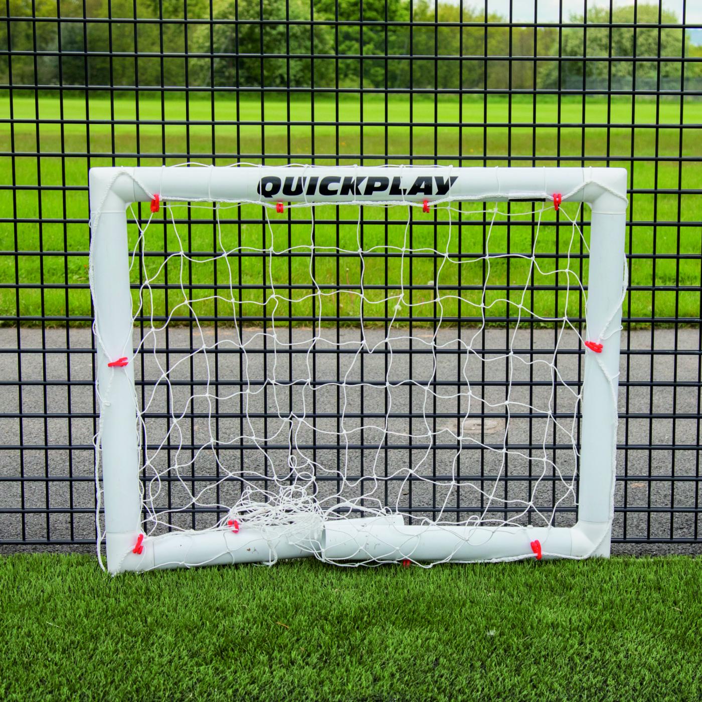 Miniportería de fútbol Q-FOLD 90 x 75 cm plegada en plano sobre césped artificial delante de una valla de malla negra.