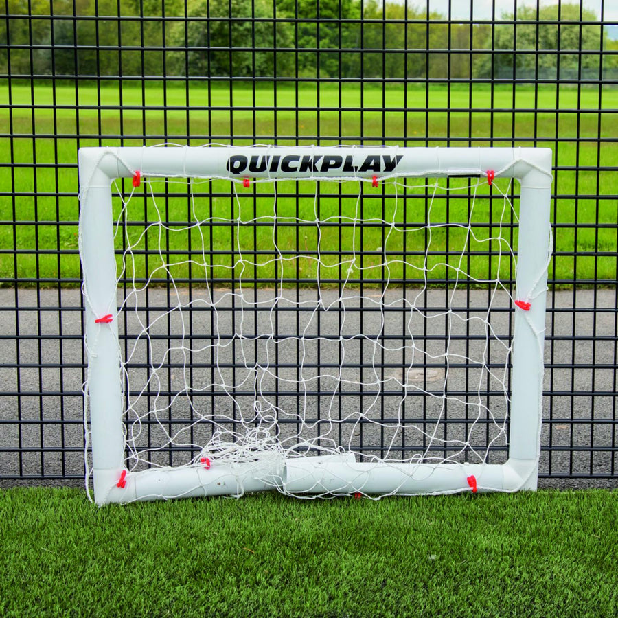 Miniportería de fútbol Q-FOLD 90 x 75 cm plegada en plano sobre césped artificial delante de una valla de malla negra.