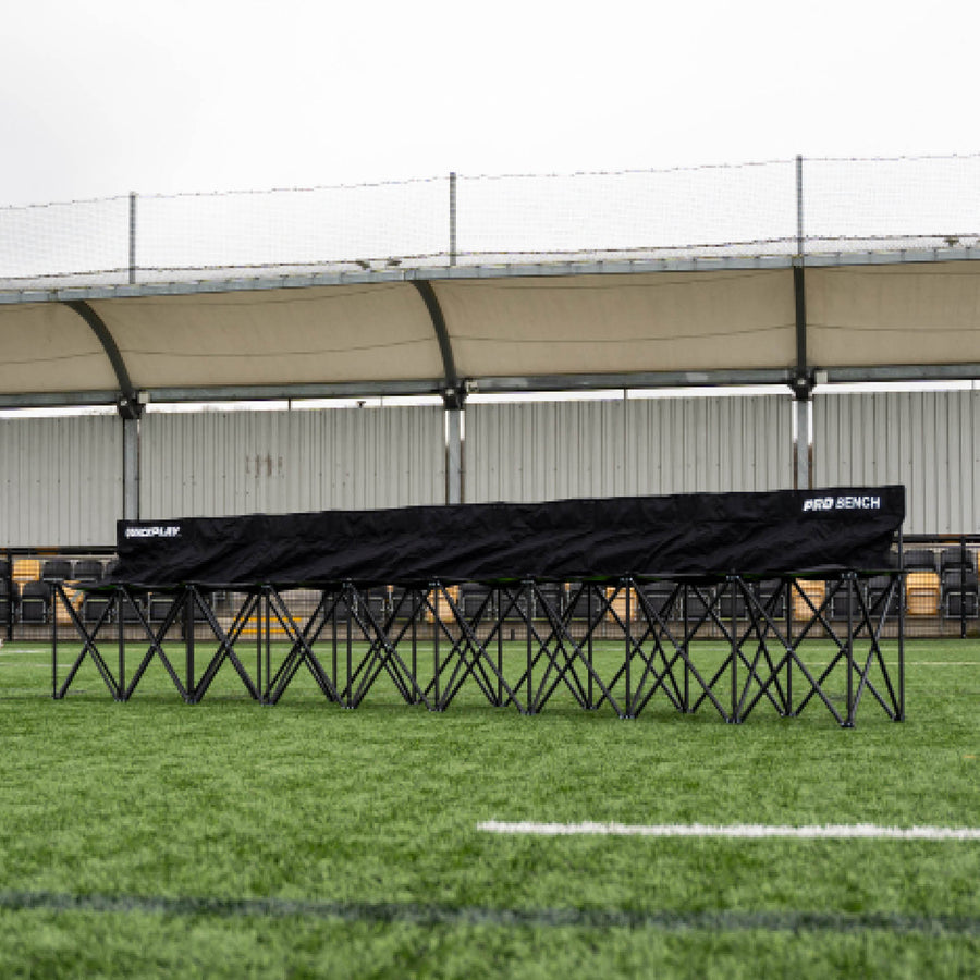 Banquillo de suplentes PRO Bench de 9 plazas, largo y negro, desplegado sobre césped artificial delante de una grada cubierta.