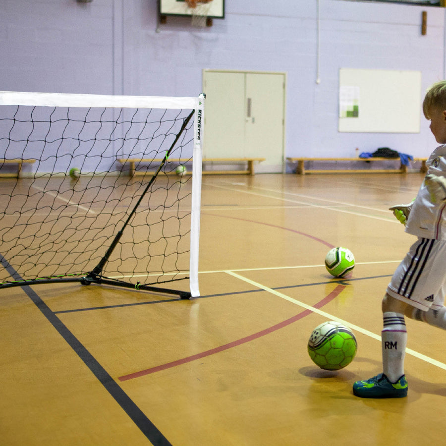 KICKSTER Elite 370 x 180 cm junto a un niño en un pabellón, a punto de chutar un balón verde a una portería pequeña.