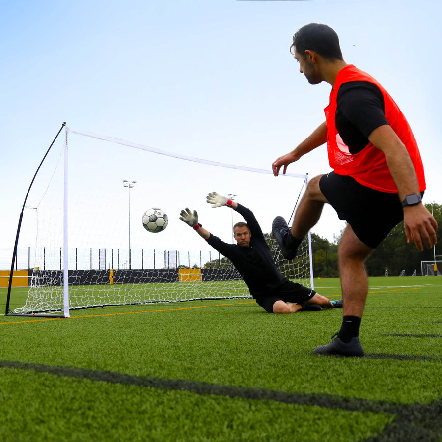 Portería KICKSTER Elite 370 x 180 cm a ras de suelo, con un portero estirándose para parar el disparo de un jugador con peto naranja.