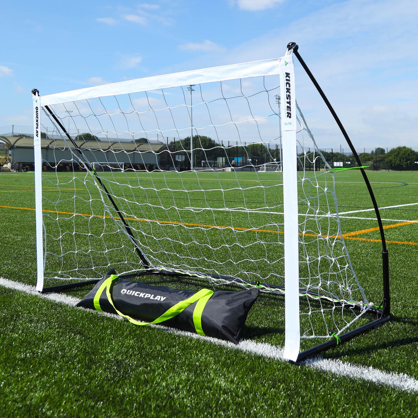 Portería KICKSTER Elite 150 x 100 cm en un campo de césped artificial, con su bolsa de transporte negra QUICKPLAY delante.