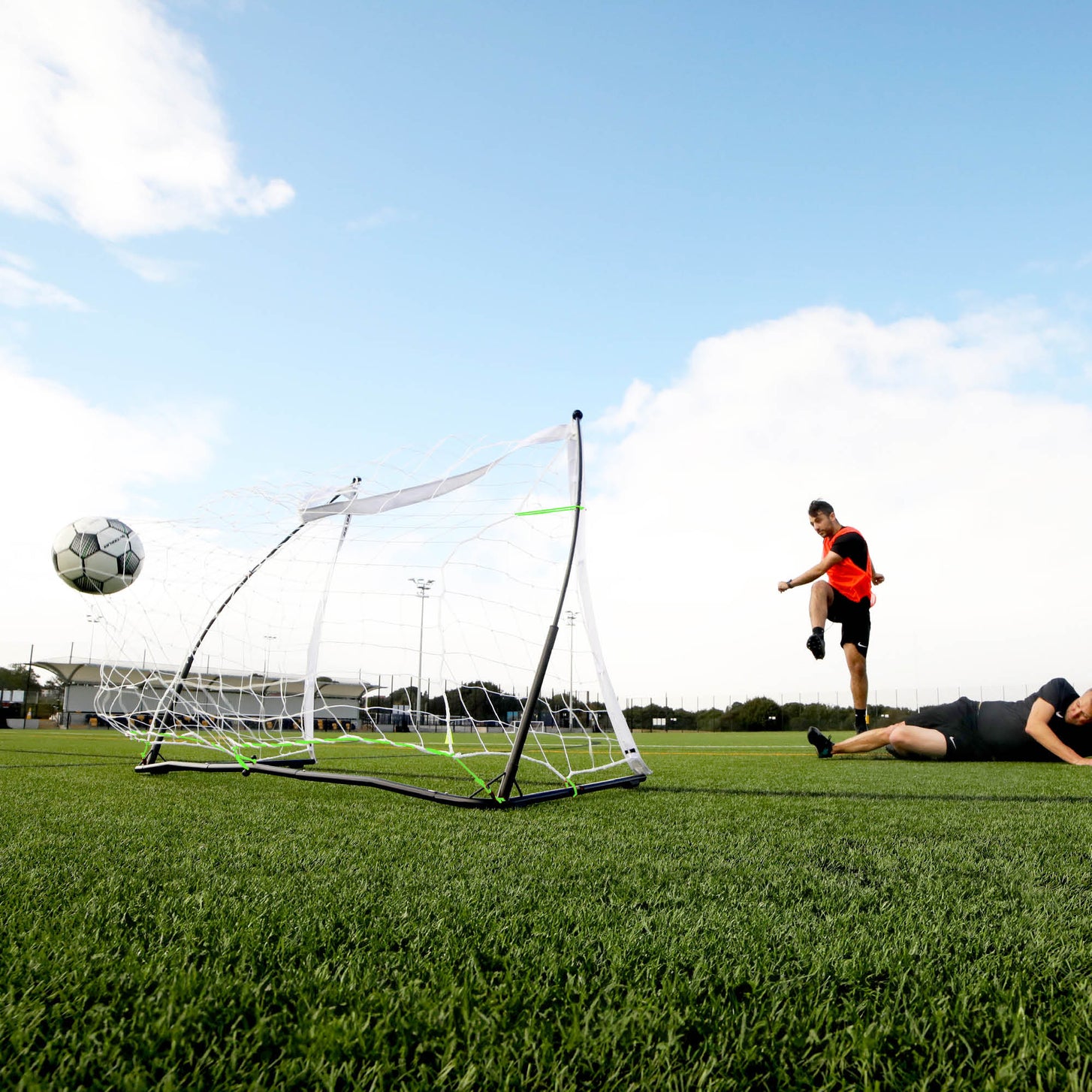 Vista desde abajo de un jugador que chuta un balón junto a la portería KICKSTER Elite 150 x 100 cm mientras otro jugador está tendido en el campo.