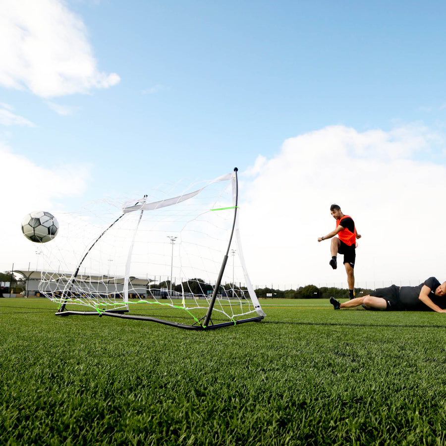 Vista desde abajo de un jugador que chuta un balón junto a la portería KICKSTER Elite 150 x 100 cm mientras otro jugador está tendido en el campo.