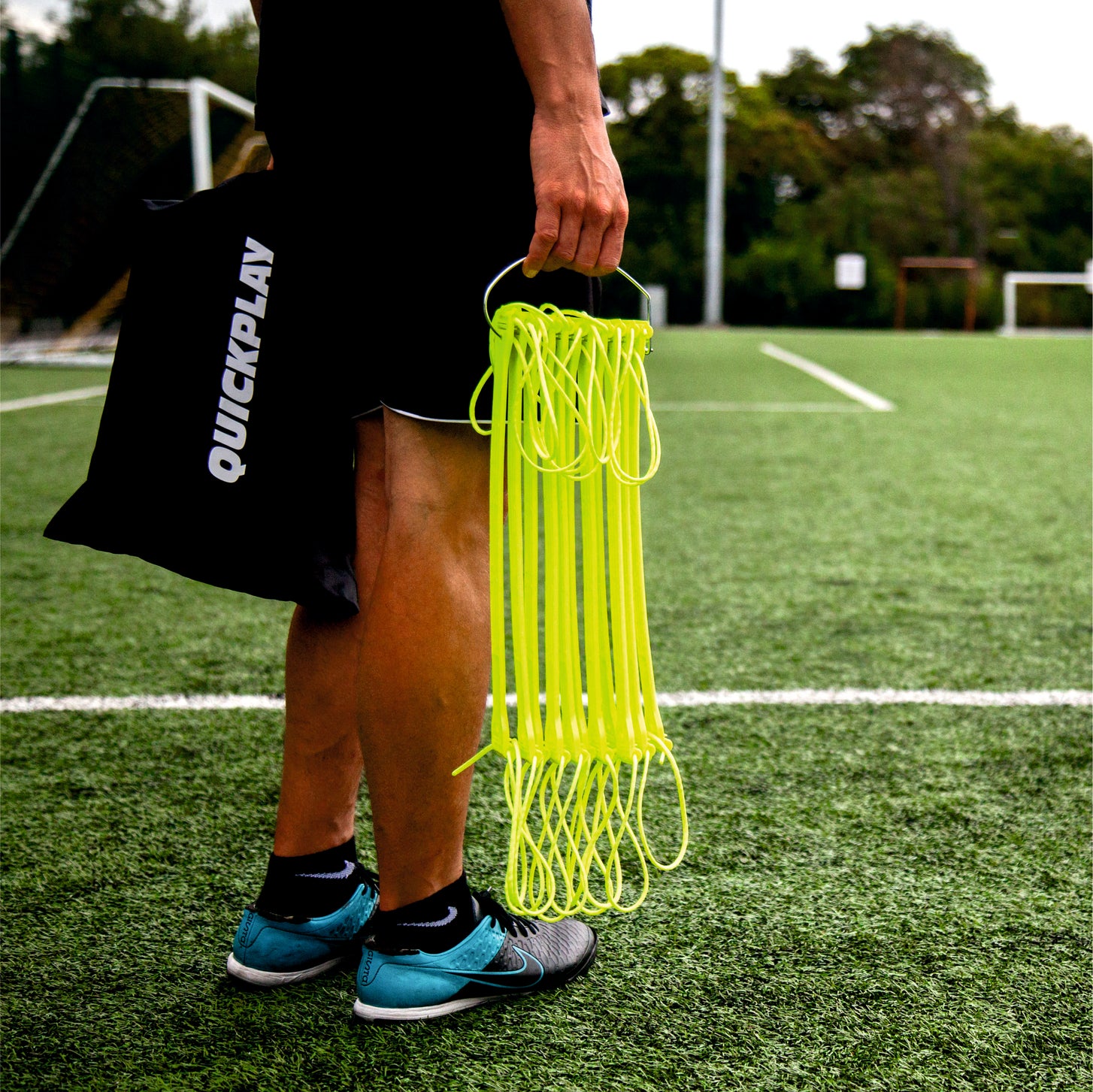 Jugador sujetando la escalera de agilidad amarilla plegada en su gancho, con una bolsa negra QUICKPLAY, en un campo de césped artificial.