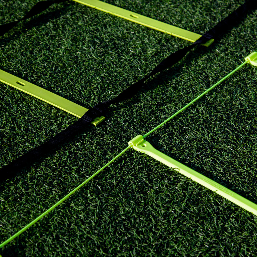 Primer plano de peldaños amarillos de la escalera de agilidad sobre césped artificial, una escalera con correas negras y otra con un fino cordón verde.
