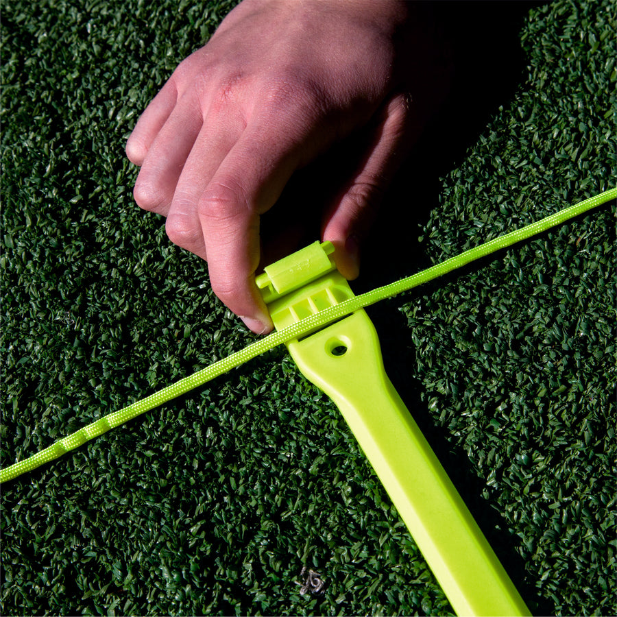 Primer plano de una mano enganchando el cordón de la escalera de agilidad en un peldaño amarillo sobre césped artificial.