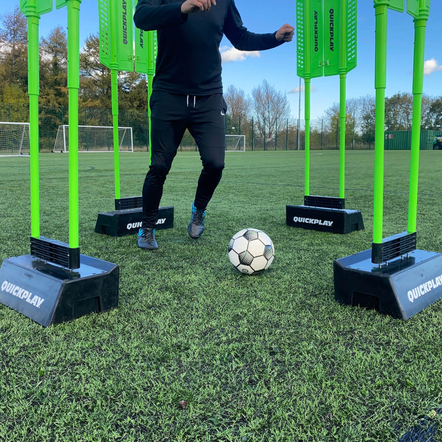 Tablas de rebote de doble ángulo REPLAY Block sosteniendo cuatro maniquíes verdes, con un jugador moviendo el balón entre ellos sobre césped artificial.