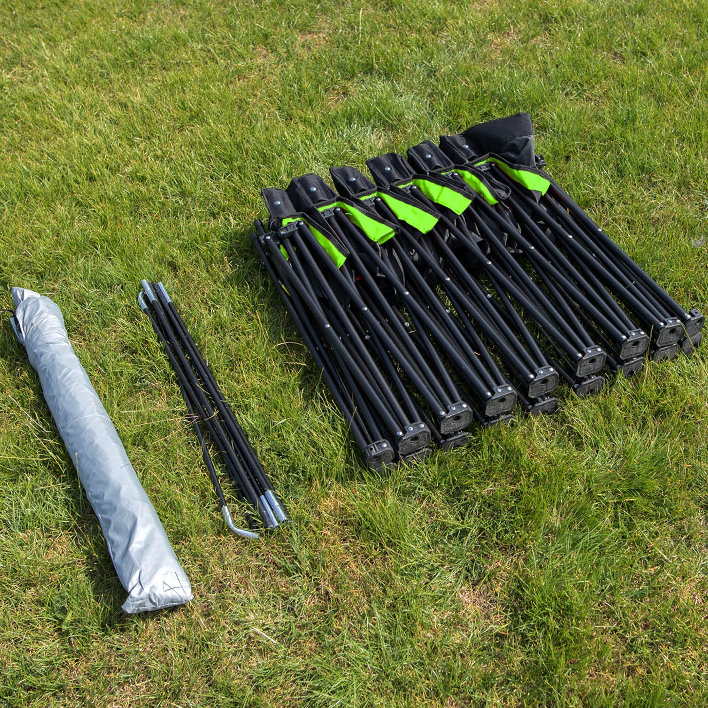 Piezas del PRO Bench Shelter dispuestas sobre la hierba: el marco del banquillo plegado, las barras del toldo y el toldo gris enrollado.