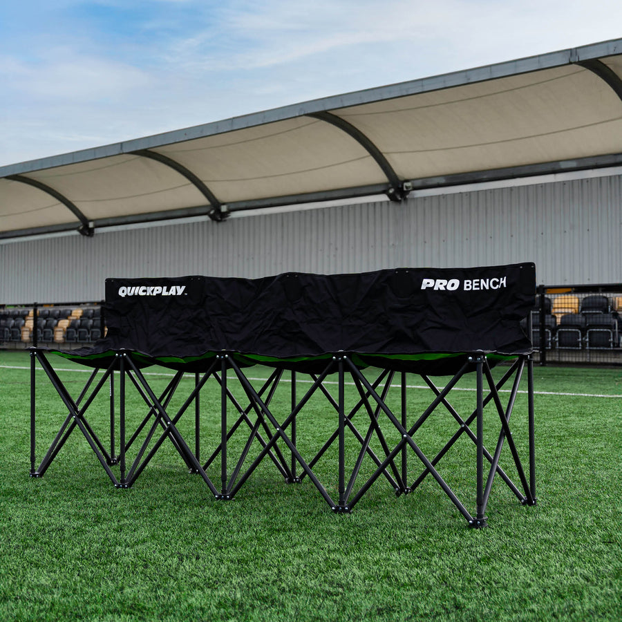 Banquillo de suplentes PRO Bench de 4 plazas, negro, desplegado sobre césped artificial delante de una grada cubierta.