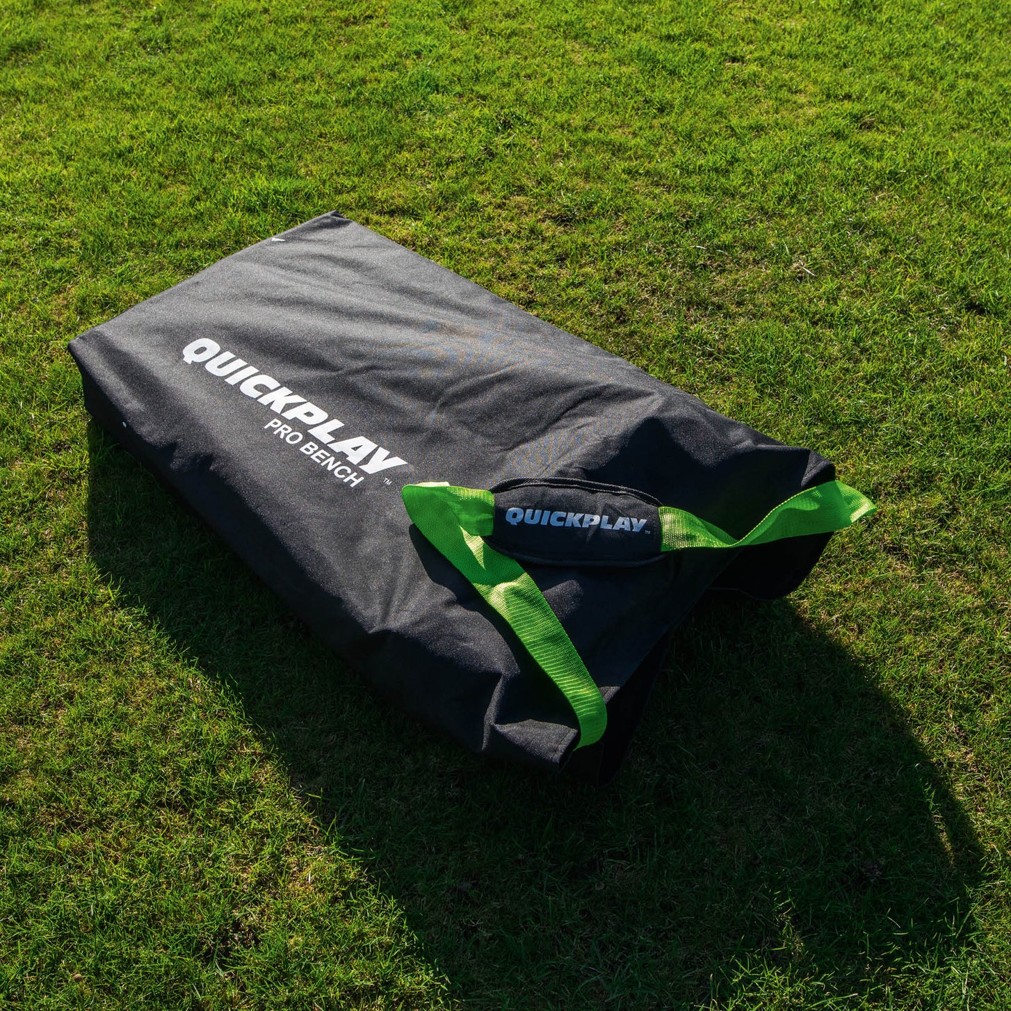 Bolsa de transporte del banquillo de suplentes PRO Bench de 4 plazas, negra con el texto QUICKPLAY PRO BENCH y correas verdes, sobre la hierba al sol.