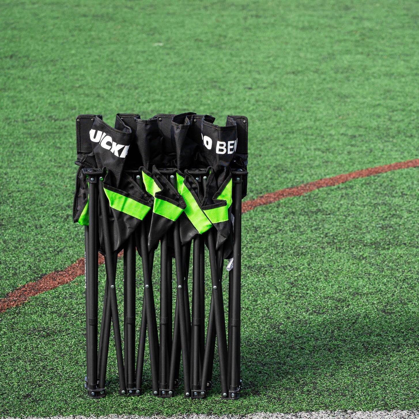 Banquillo de suplentes PRO Bench de 4 plazas plegado en vertical sobre césped artificial, con los asientos negros y las correas verdes recogidos.