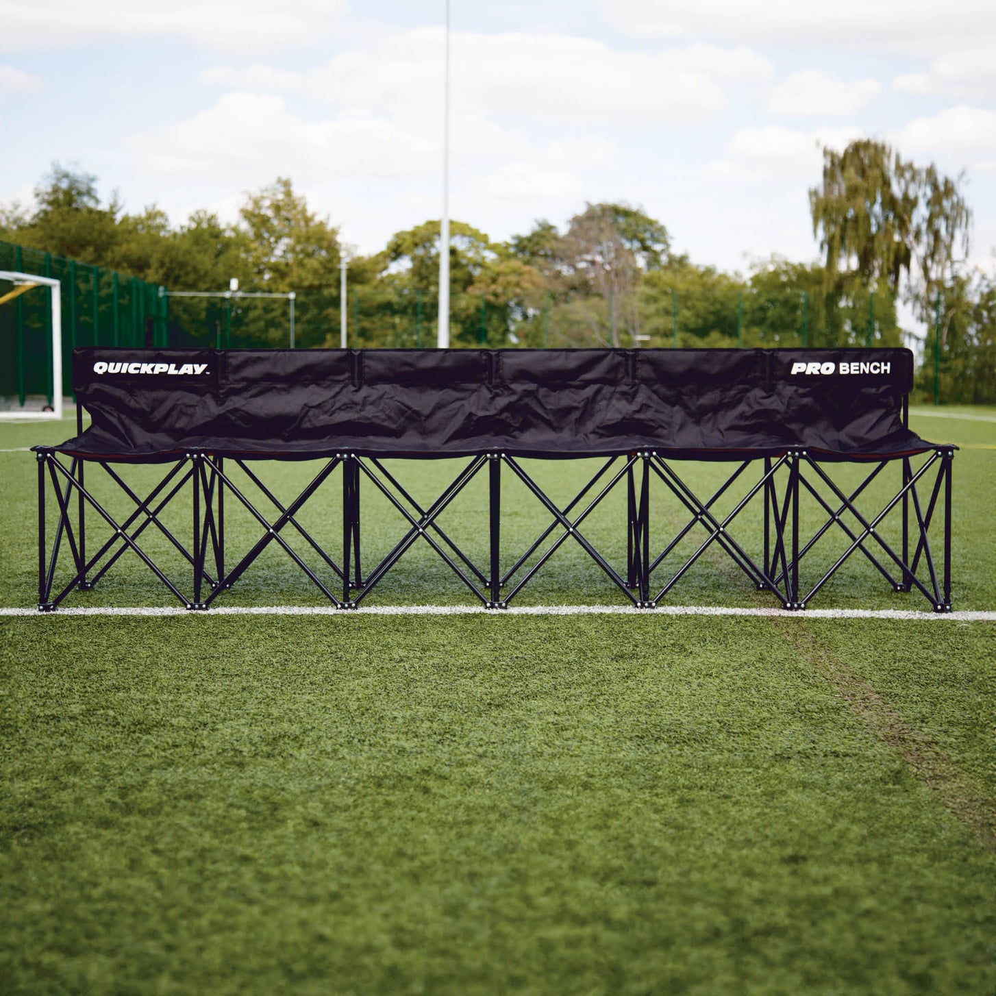 Banquillo de suplentes PRO Bench de 6 plazas desplegado a lo largo de la banda de un campo de hierba, vista frontal con árboles detrás.