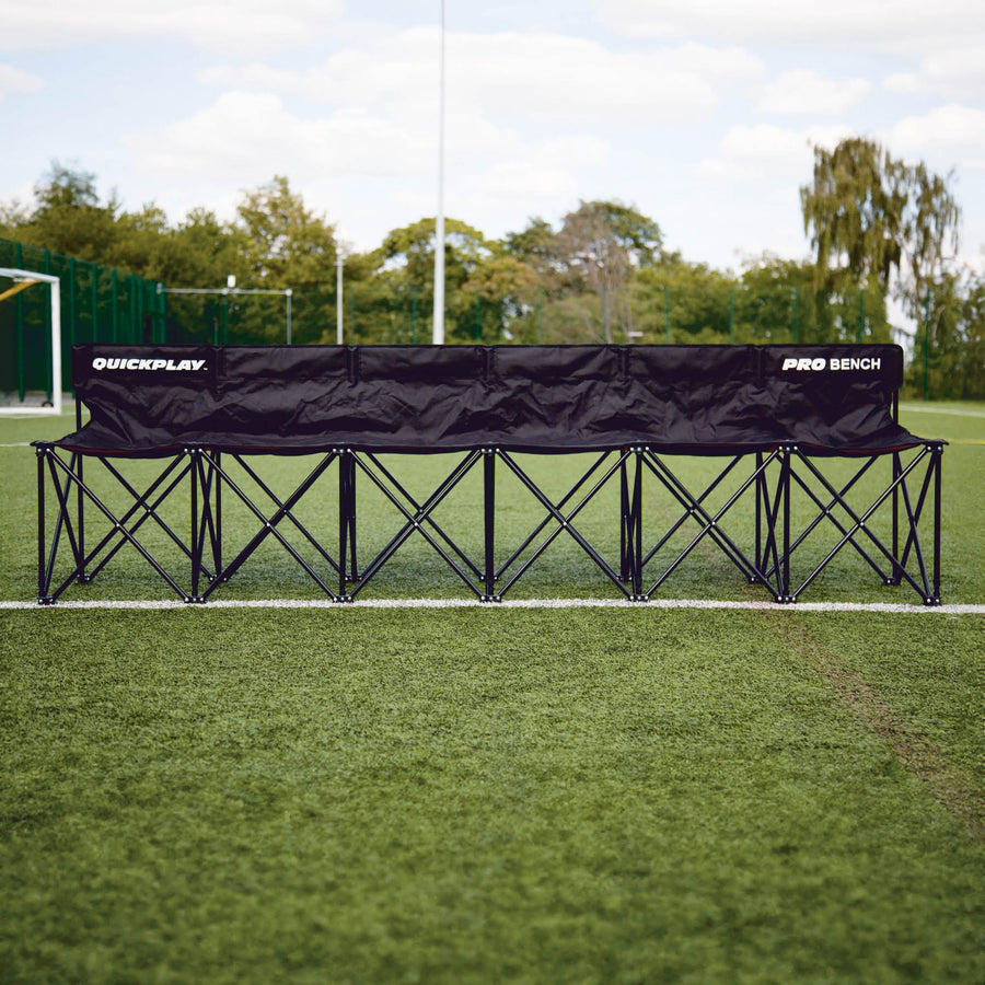 Banquillo de suplentes PRO Bench de 6 plazas desplegado a lo largo de la banda de un campo de hierba, vista frontal con árboles detrás.