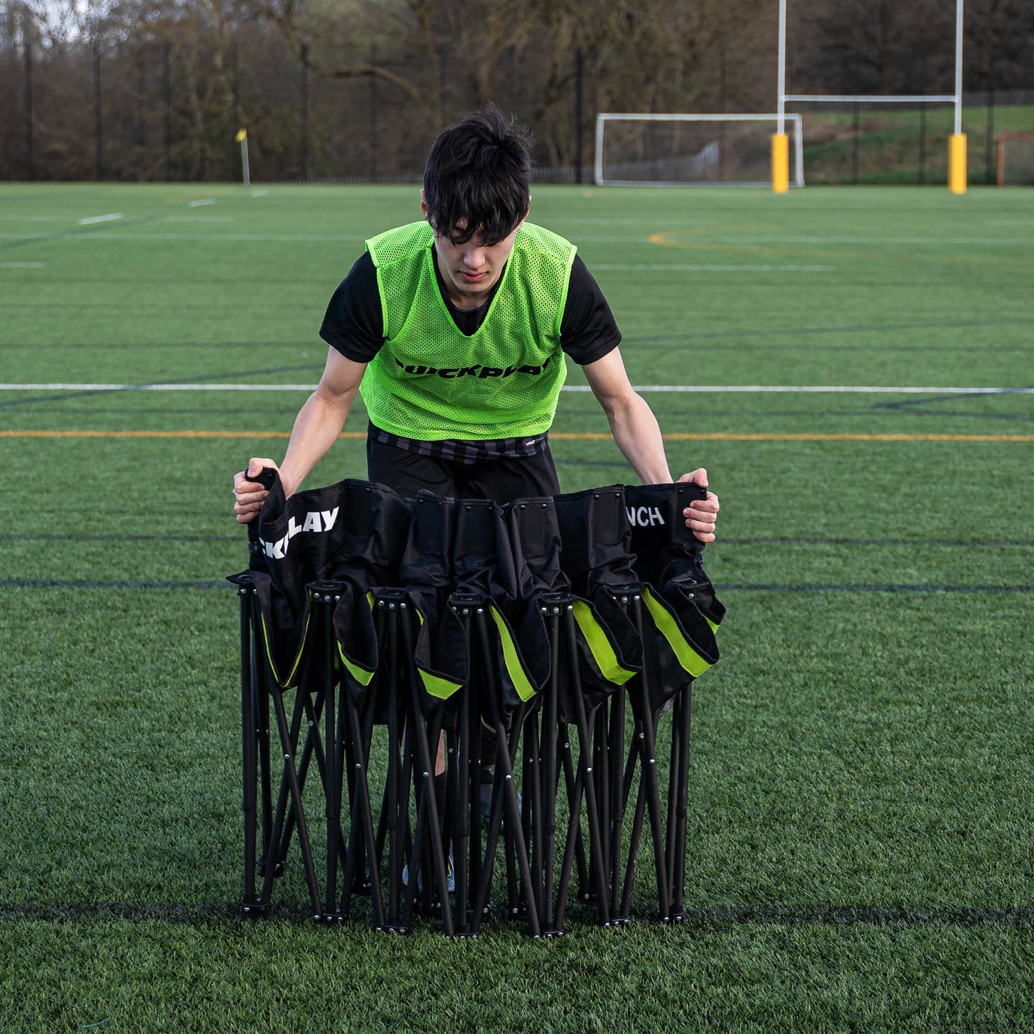 Jugador con un peto verde QUICKPLAY sujetando el banquillo de suplentes PRO Bench de 6 plazas a medio plegar en un campo de césped artificial.