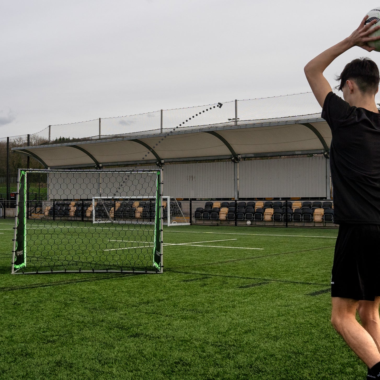 Jugador sacando de banda hacia el TEKKERS GS Trainer en un campo de césped artificial, con una línea punteada que sigue el balón.