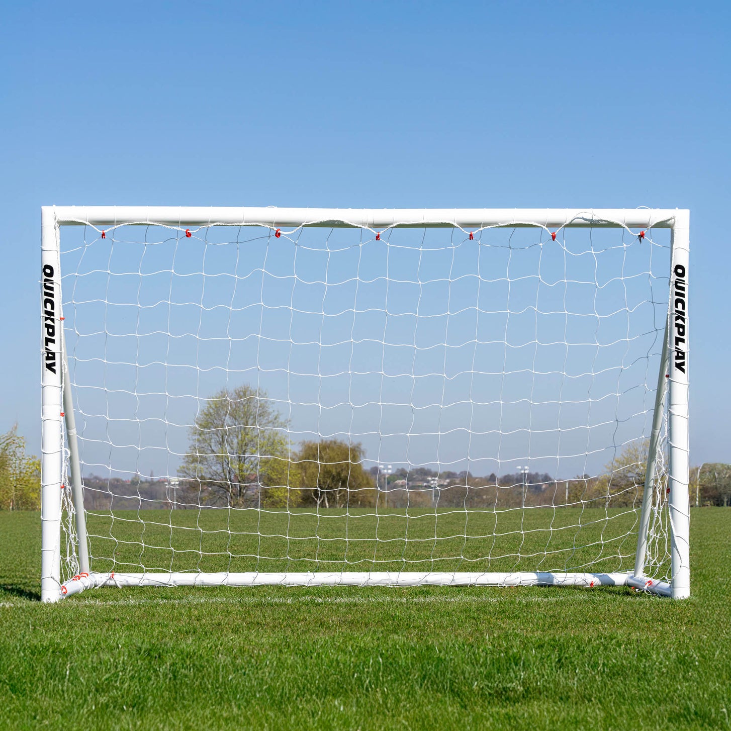 Vista frontal de la portería de fútbol Q-FOLD 240 x 150 cm en un campo de hierba, con árboles visibles a través de la red blanca.