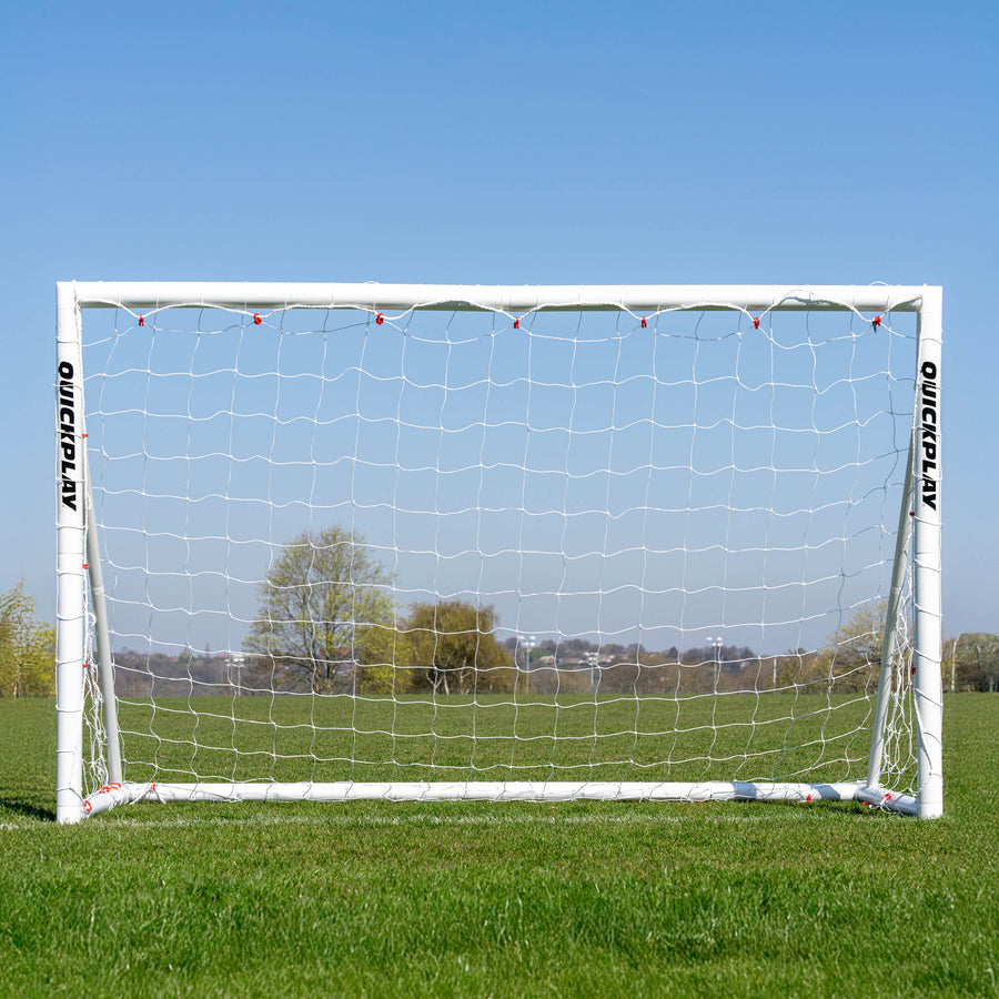 Vista frontal de la portería de fútbol Q-FOLD 240 x 150 cm en un campo de hierba, con árboles visibles a través de la red blanca.