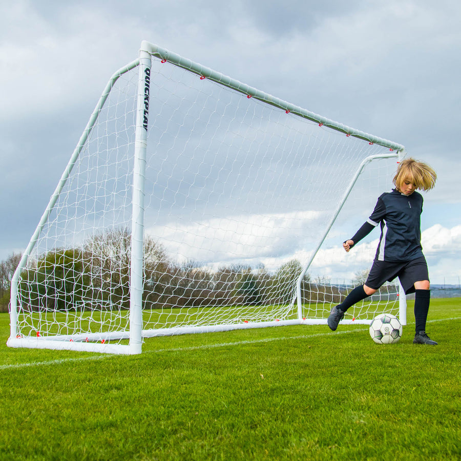 Niño regateando con un balón junto a la portería Q-FOLD Match 370 x 180 cm en un campo de hierba bajo un cielo nublado.
