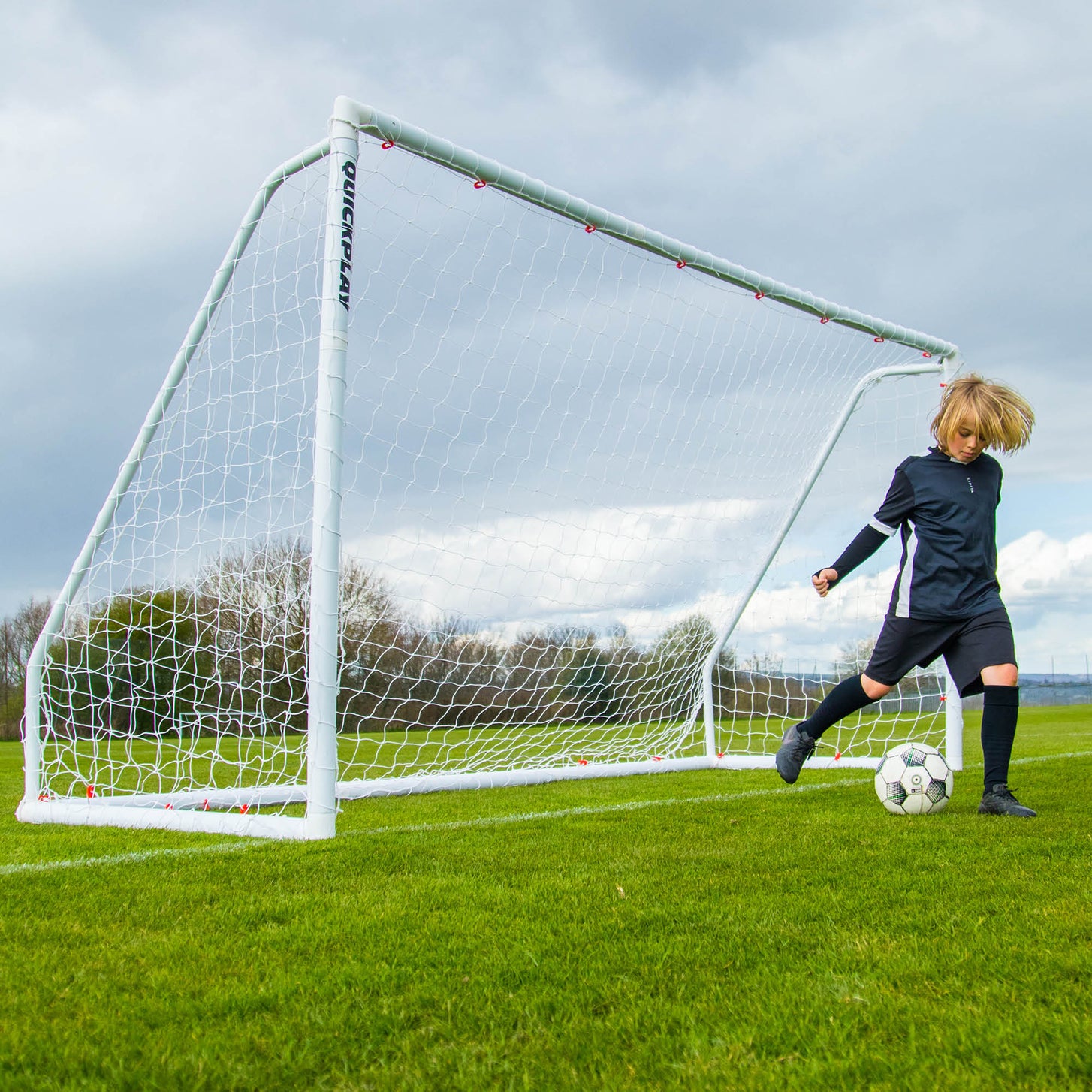 Niño regateando con un balón delante de una portería blanca Q-FOLD Match en un campo de hierba bajo un cielo nublado.