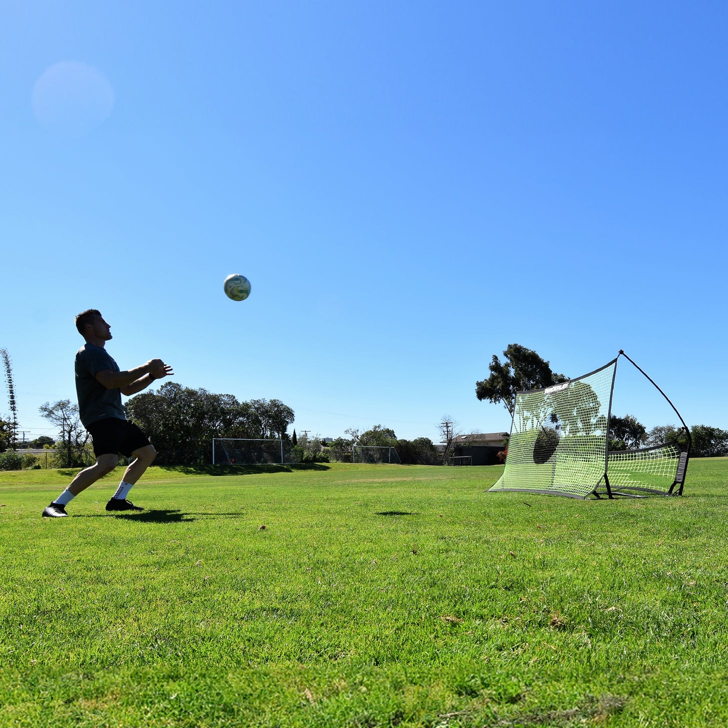 Vista amplia de un jugador preparándose para controlar un balón en el aire, con el rebotador TEKKERS Trainer detrás de él sobre la hierba.