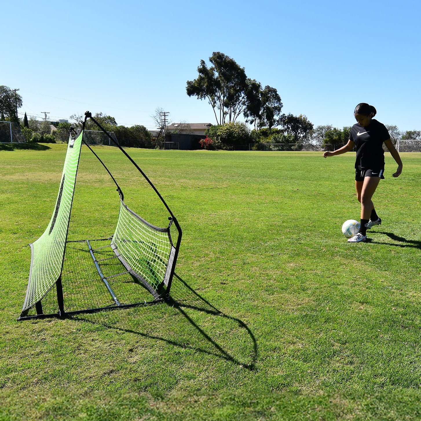 Jugador a punto de pasar un balón al rebotador de fútbol TEKKERS Trainer con doble red, visto de lado sobre hierba.