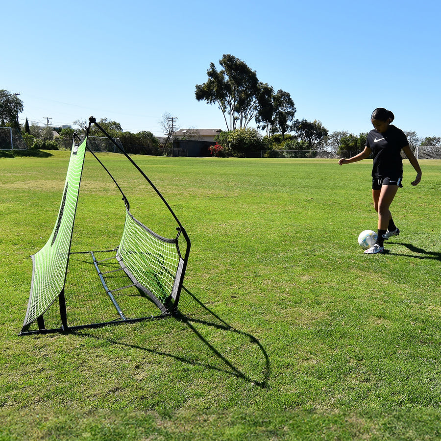 Jugador a punto de pasar un balón al rebotador de fútbol TEKKERS Trainer con doble red, visto de lado sobre hierba.