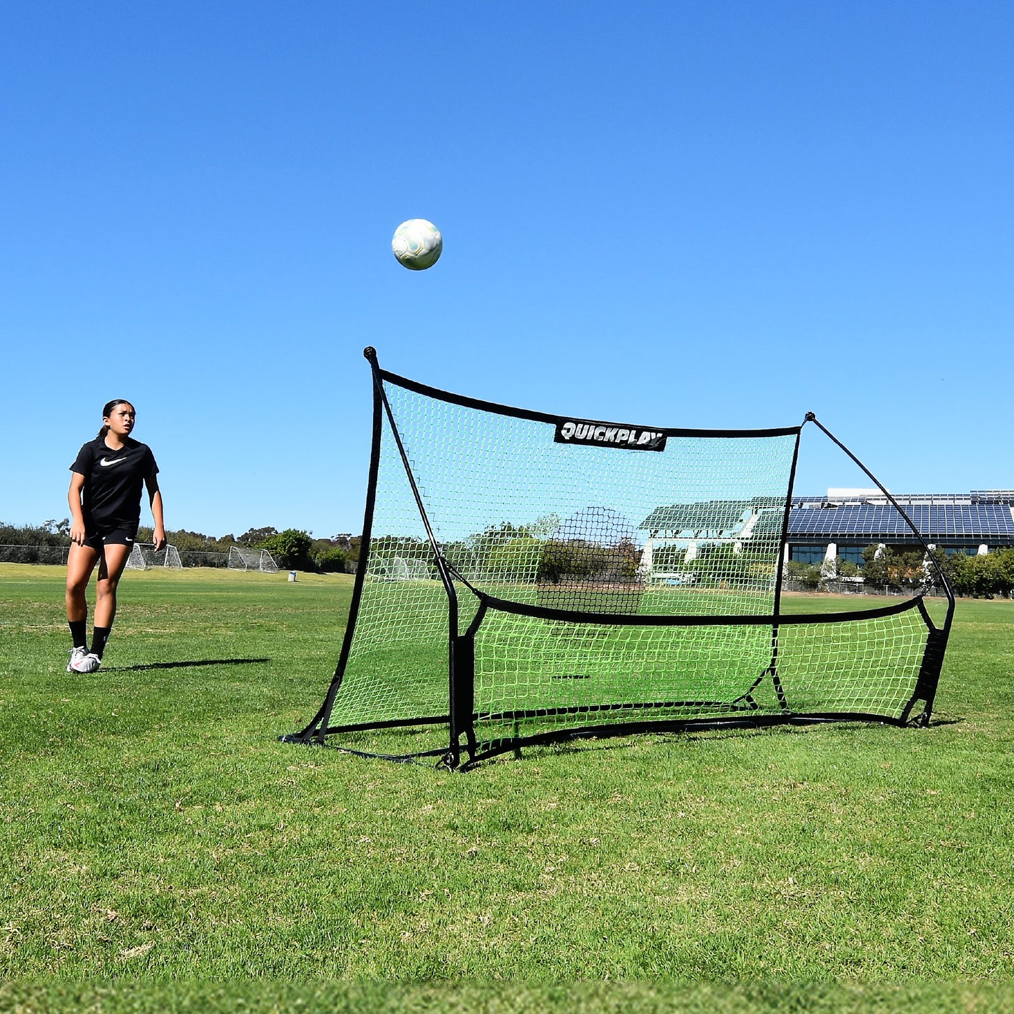 Jugador mirando cómo sube un balón desde la red del TEKKERS Trainer, vista frontal del rebotador en un campo de hierba.
