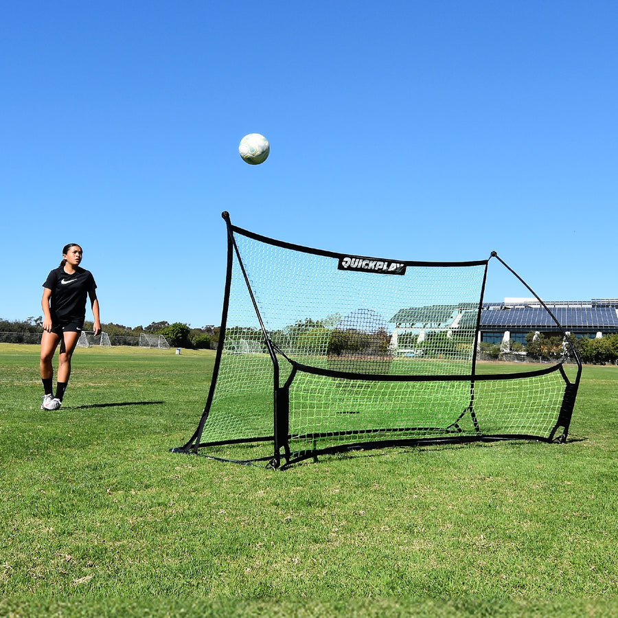 Jugador mirando cómo sube un balón desde la red del TEKKERS Trainer, vista frontal del rebotador en un campo de hierba.